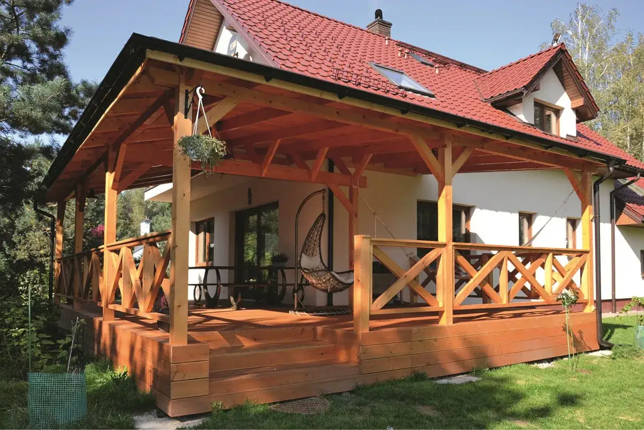 Drewniany taras zadaszony, z balustradą i schodami. Widoczne drzwi tarasowe otwierane na zewnątrz, wiszący fotel i stoły.