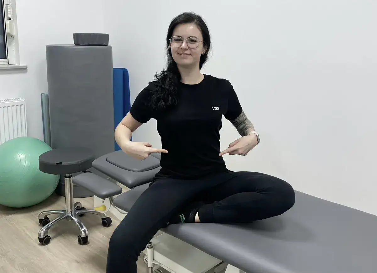 Kobieta pokazuje, jak sprawdzić rozejście mięśnia prostego brzucha. W tle sprzęt rehabilitacyjny.