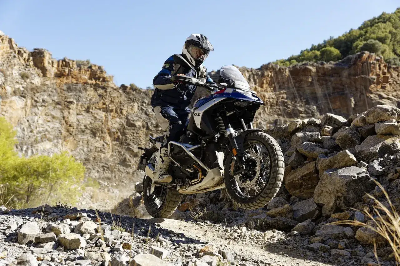 Motocyklista na motocyklu typu adv pokonuje kamienisty teren, unosząc się w powietrzu.