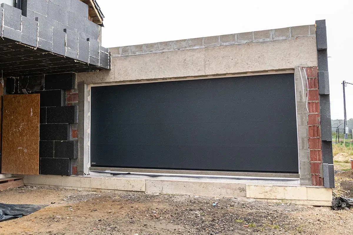 Montaż bramy garażowej w trakcie budowy. Ciemnoszara brama segmentowa w otworze, z widocznymi elementami izolacji i konstrukcji budynku.