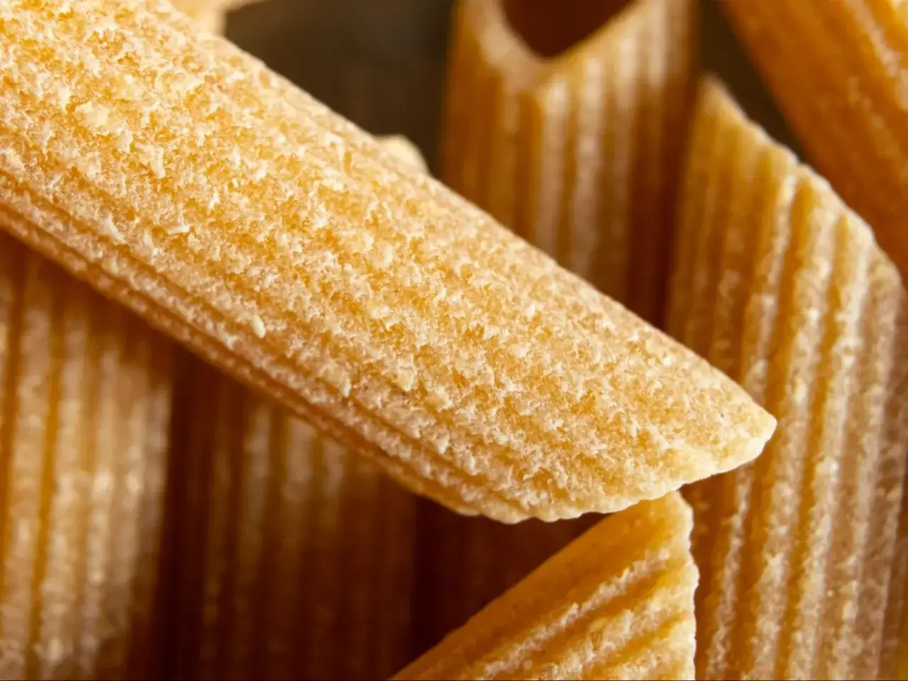 Zbliżenie na jasny makaron penne, z widocznymi rowkami. Kontrastuje z ciemniejszym tłem.