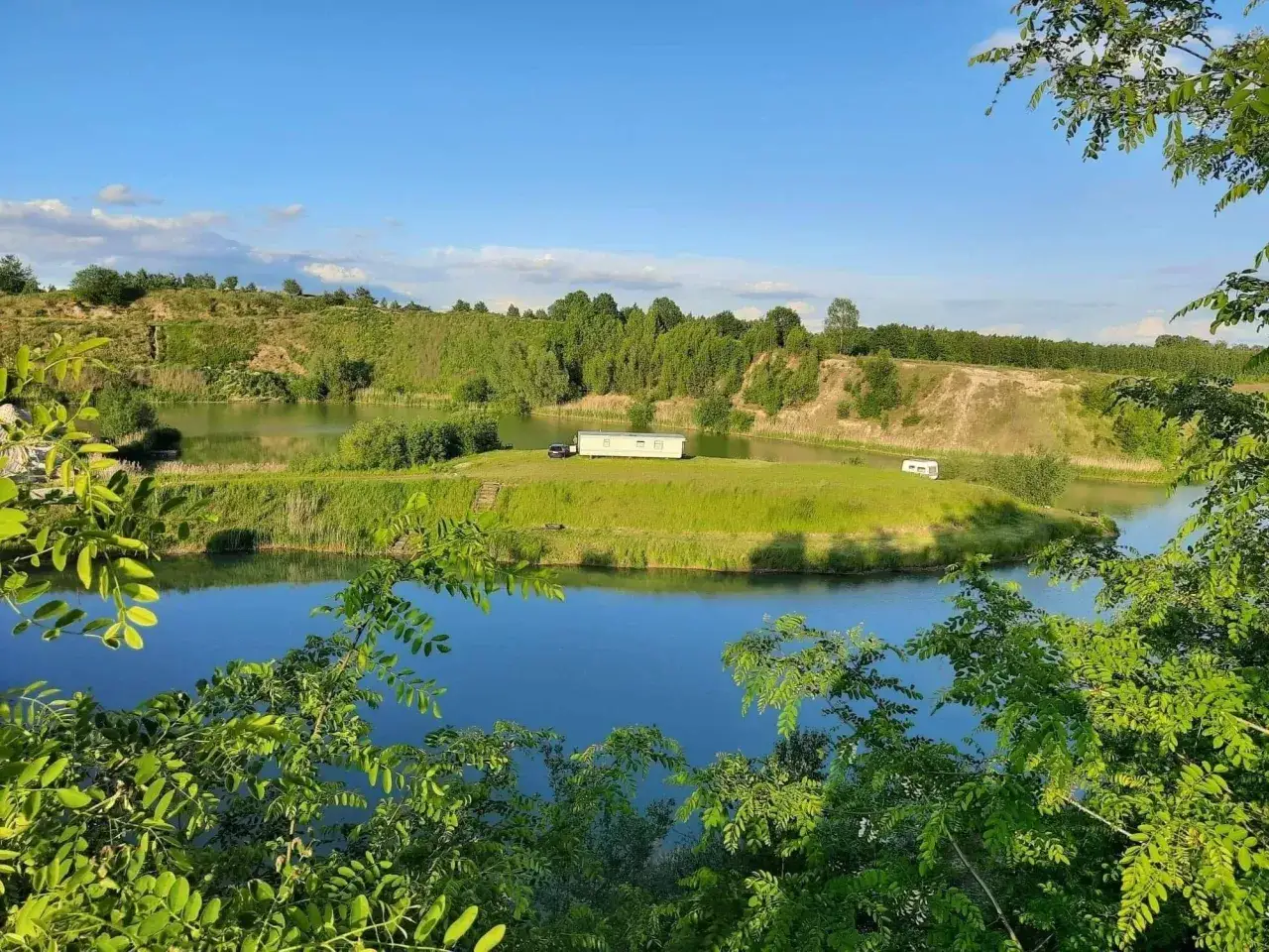 Zielona wyspa na spokojnym łowisku, z przyczepą kempingową i domkiem, otoczona drzewami i błękitnym niebem.