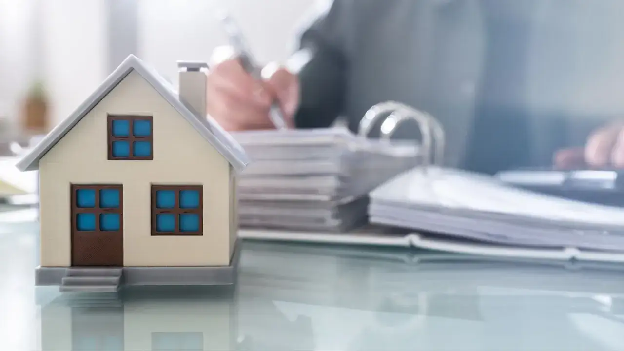 Model domu na biurku, obok stosów dokumentów. Czy kupując dom od dewelopera płacę podatek? Analiza formalności.