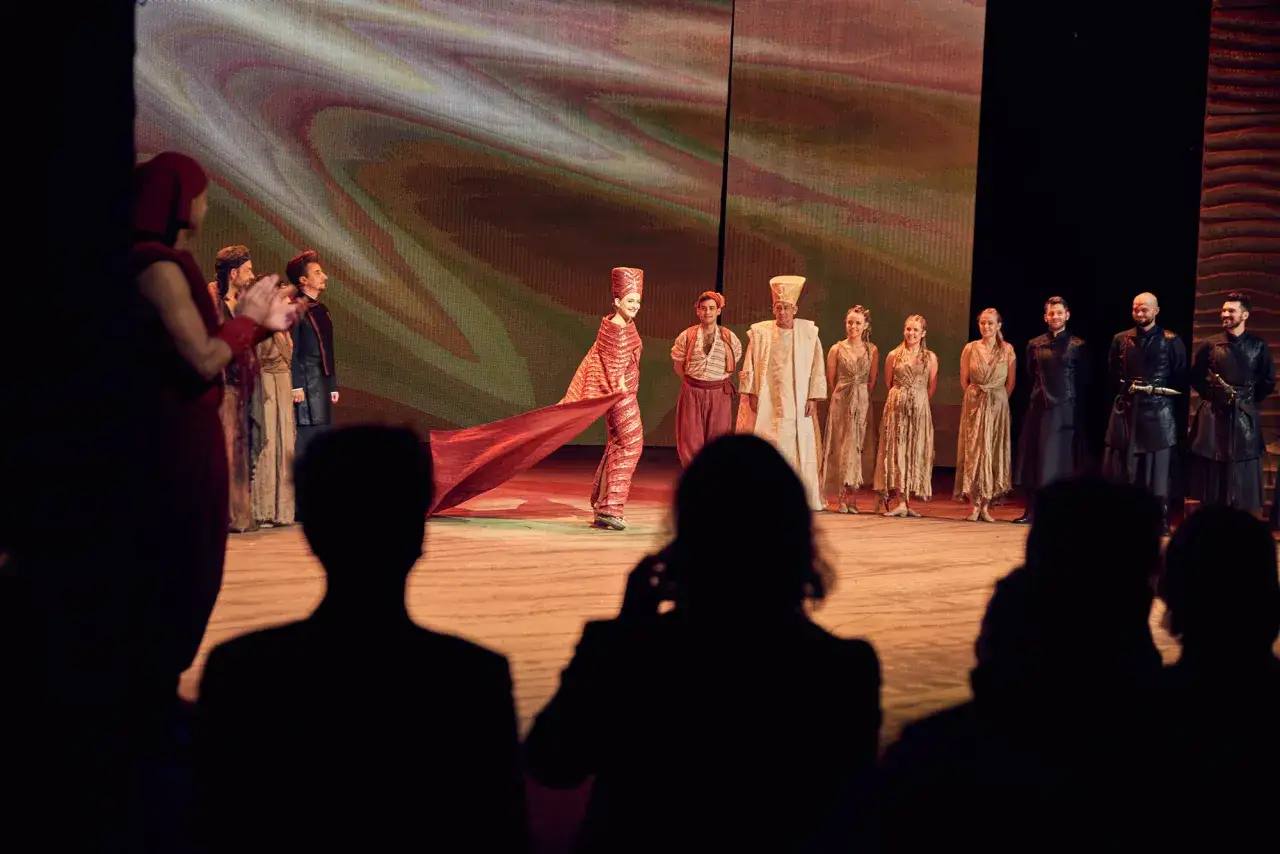 Aida w Teatrze Roma. Aktorka w czerwonej sukni z trenem, obok niej inni artyści w strojach z epoki. Spektakl trwa, widzowie czekają na rozw&oacute;j akcji.