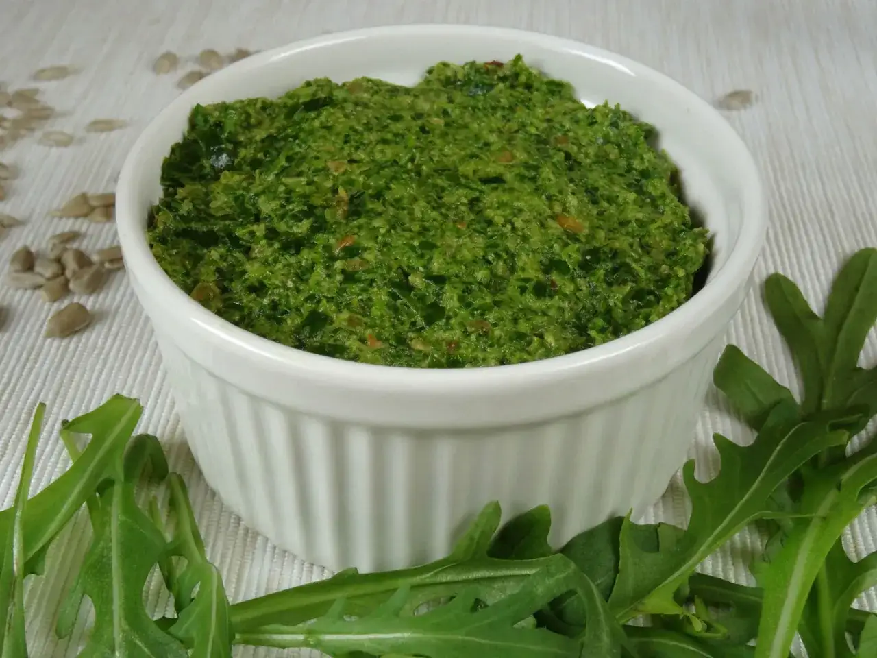 Pesto z rukoli w białej miseczce, otoczone świeżymi liśćmi rukoli i nasionami słonecznika.