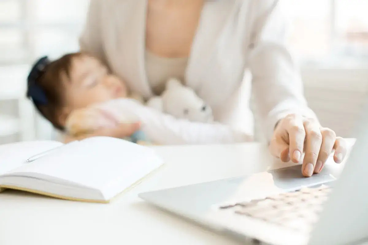 Kobieta z dzieckiem na rękach szuka informacji, jak zatrudnić nianię, korzystając z laptopa. Obok leży otwarty notatnik.