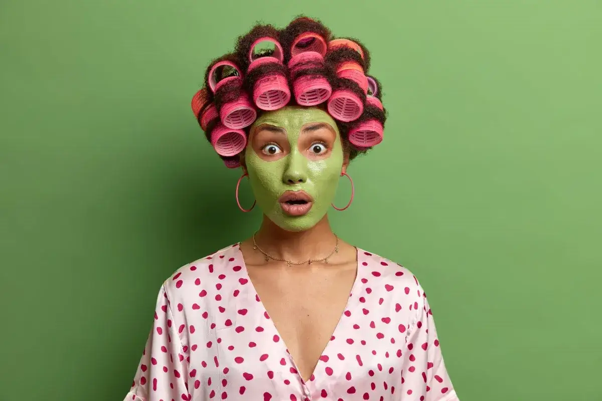 Zaskoczona kobieta z zieloną maseczką na twarzy i wałkami na włosach. Dowiedz się, z czym nie łączyć witaminy C na twarz, by uniknąć podrażnień.