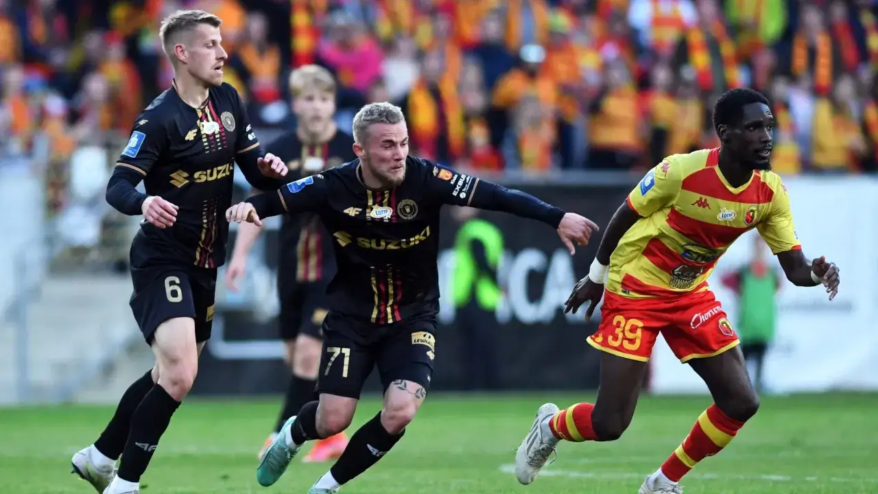 Piłkarze w czarnych strojach walczą o utrzymanie w lidze, podczas gdy zawodnik w żółto-czerwonym trykocie biegnie w stronę bramki, być może decydując, kto spada z ekstraklasy.