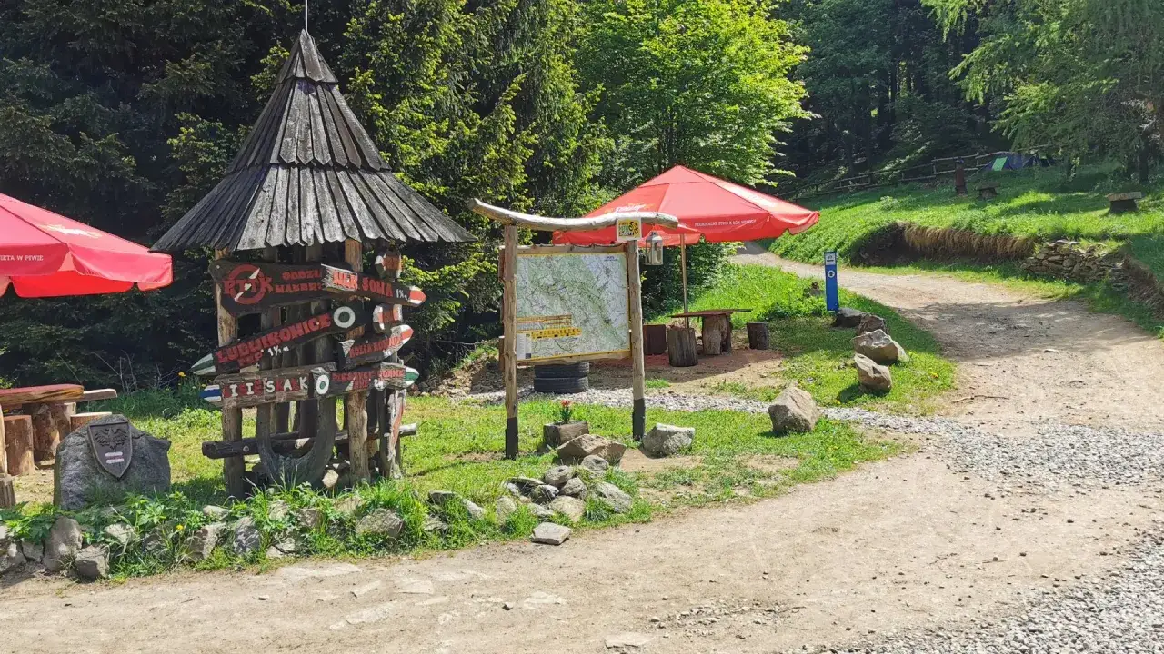 Drewniany drogowskaz przy szlaku w Górach Sowich. Obok mapa, czerwone parasole i ławki.