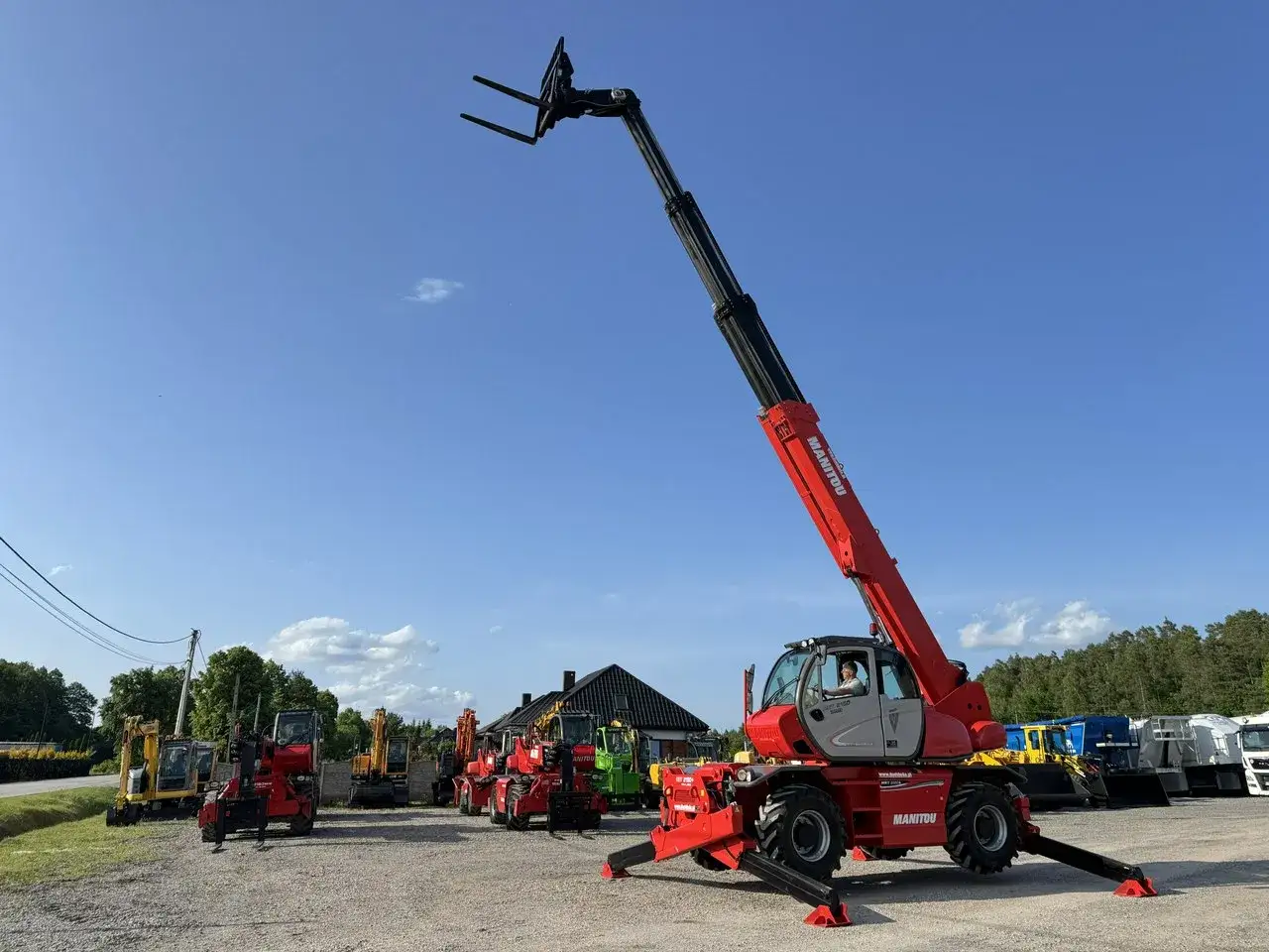 Manitou MRT 2150 dane techniczne: czerwony ładowarka teleskopowa z wysuniętym wysięgnikiem i widłami, stabilizatory rozłożone. W tle inne maszyny budowlane.