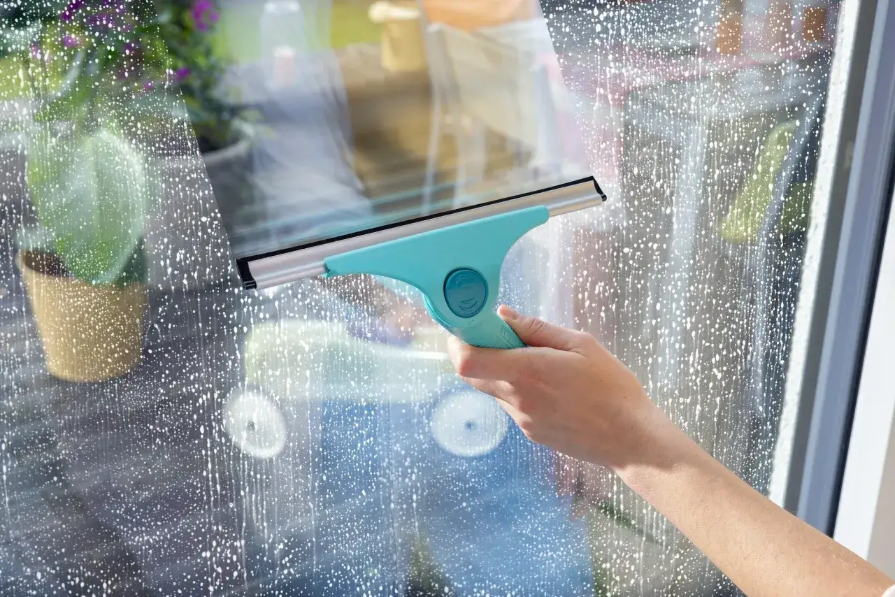 Ręka trzyma turkusową ściągaczkę, pokazując, jak myć okna, usuwając pianę i smugi.