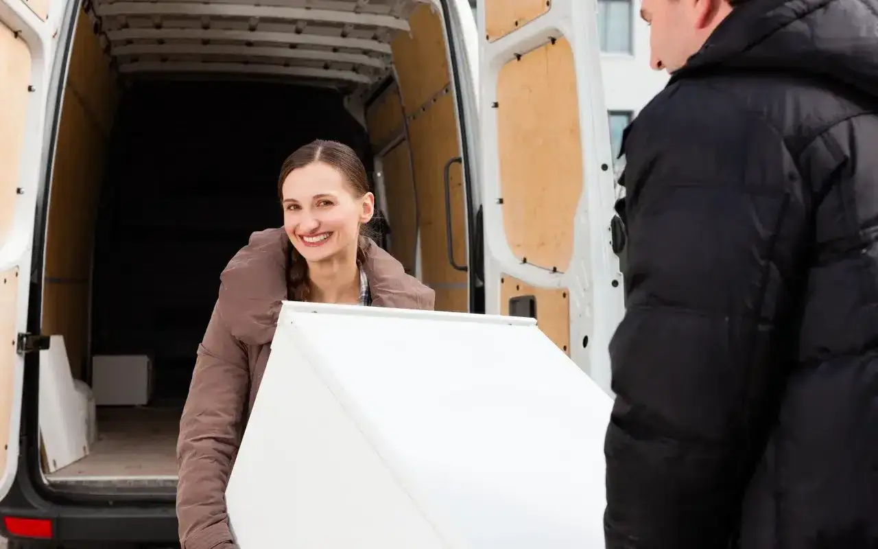 Uśmiechnięta kobieta i mężczyzna przenoszą białą lod&oacute;wkę do furgonetki. To praktyczny przykład, jak przewozić lod&oacute;wkę.