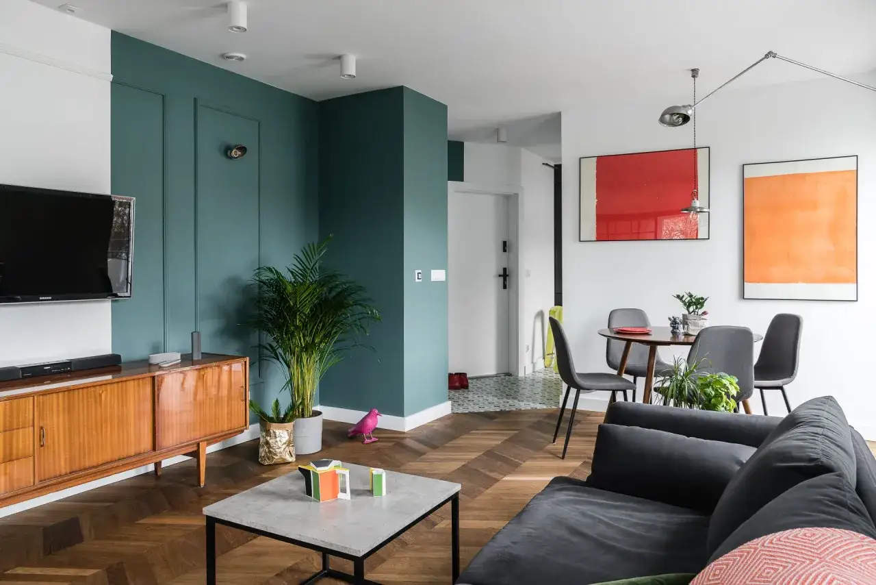 Nowoczesne mieszkanie w stylu PRL z drewnianą komodą, zieloną ścianą i kolorowymi akcentami.