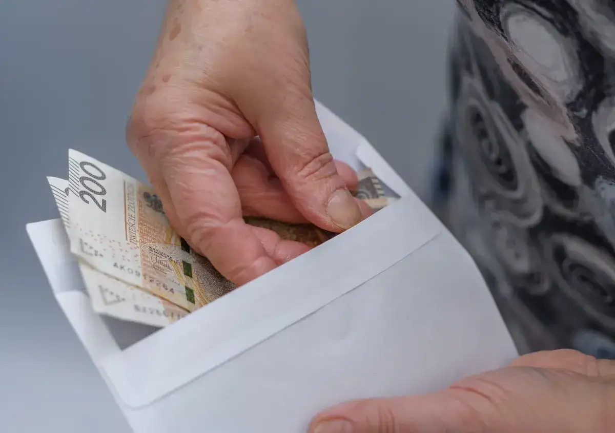 Dłoń wkłada banknoty 200 zł do koperty. Może to być związane ze złożeniem wniosku o zniżki na prąd dla emeryt&oacute;w.