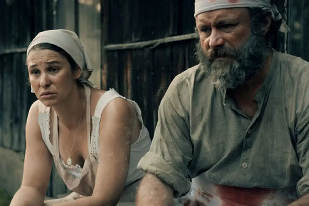 Kobieta i mężczyzna w strojach z epoki, siedzący przed drewnianą chatą. To właśnie tutaj rozgrywa się akcja filmu 