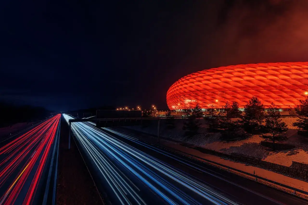 Nocna fotografia autostrady z czerwonymi i niebieskimi smugami świateł samochodowych obok stadionu oświetlonego na czerwono.
