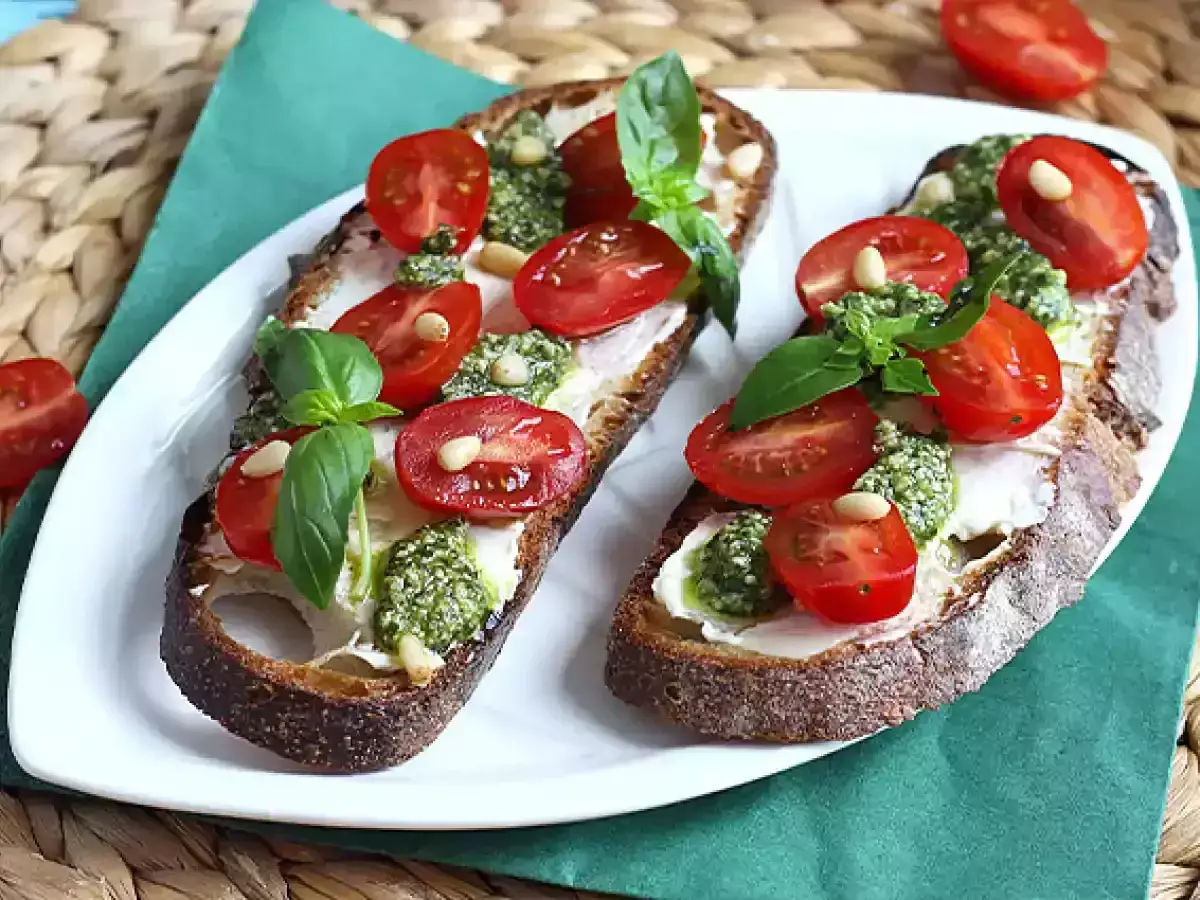 Dwa tosty z ciemnego pieczywa z kremowym serkiem, aromatycznym pesto na kanapki, pomidorkami koktajlowymi i orzeszkami pinii.