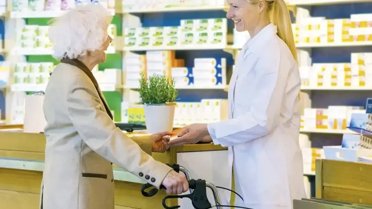 Starsza pani z balkonikiem odbiera lek od farmaceutki. Czy leki refundowane wszędzie kosztują tyle samo?