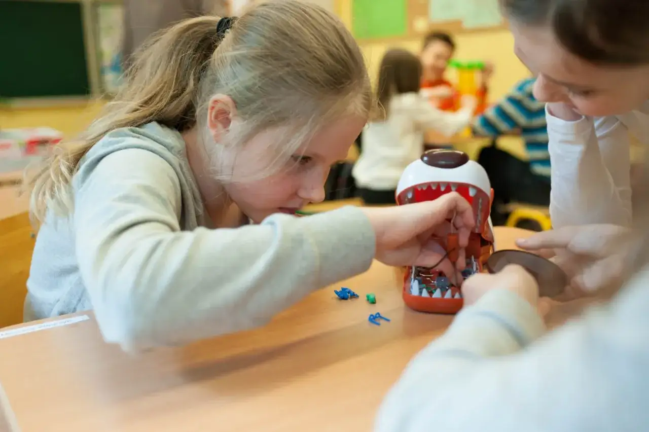 Dziewczynka z blond warkoczem skupiona na zadaniach dla 10 latka, naprawia zabawkę-potwora. Inne dzieci pracują w tle.