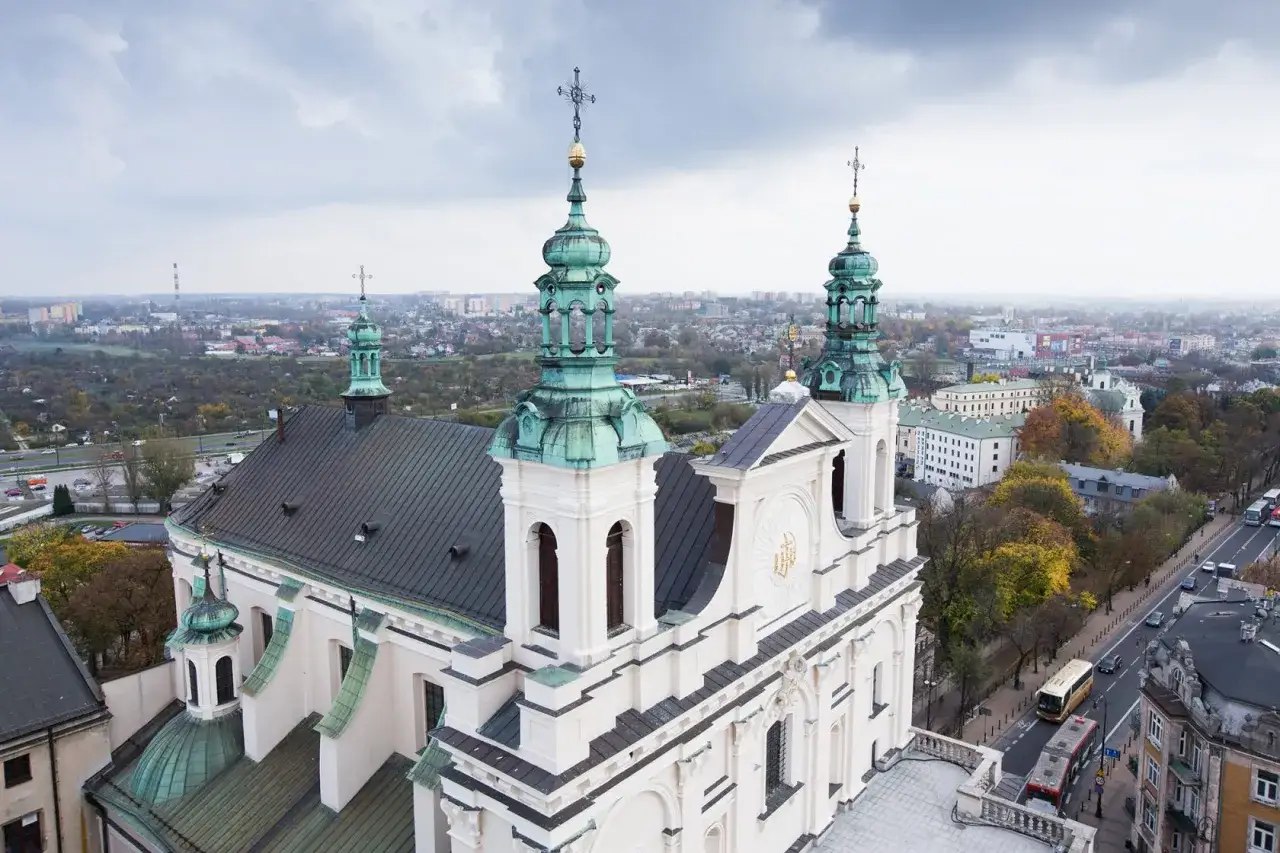 Widok z lotu ptaka na kościół z zielonymi kopułami i wieżami, rozciągający się nad miastem. W tle widać zabudowania i drzewa, a po prawej stronie ulicę z ruchem miejskim. To malowniczy fragment lublin stare miasto.