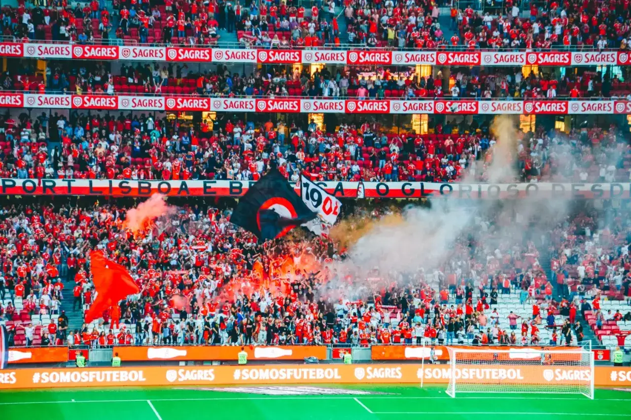 Tłum kibiców klubu piłkarskiego z Lizbony, odpalone race i flagi tworzą gorącą atmosferę na stadionie.