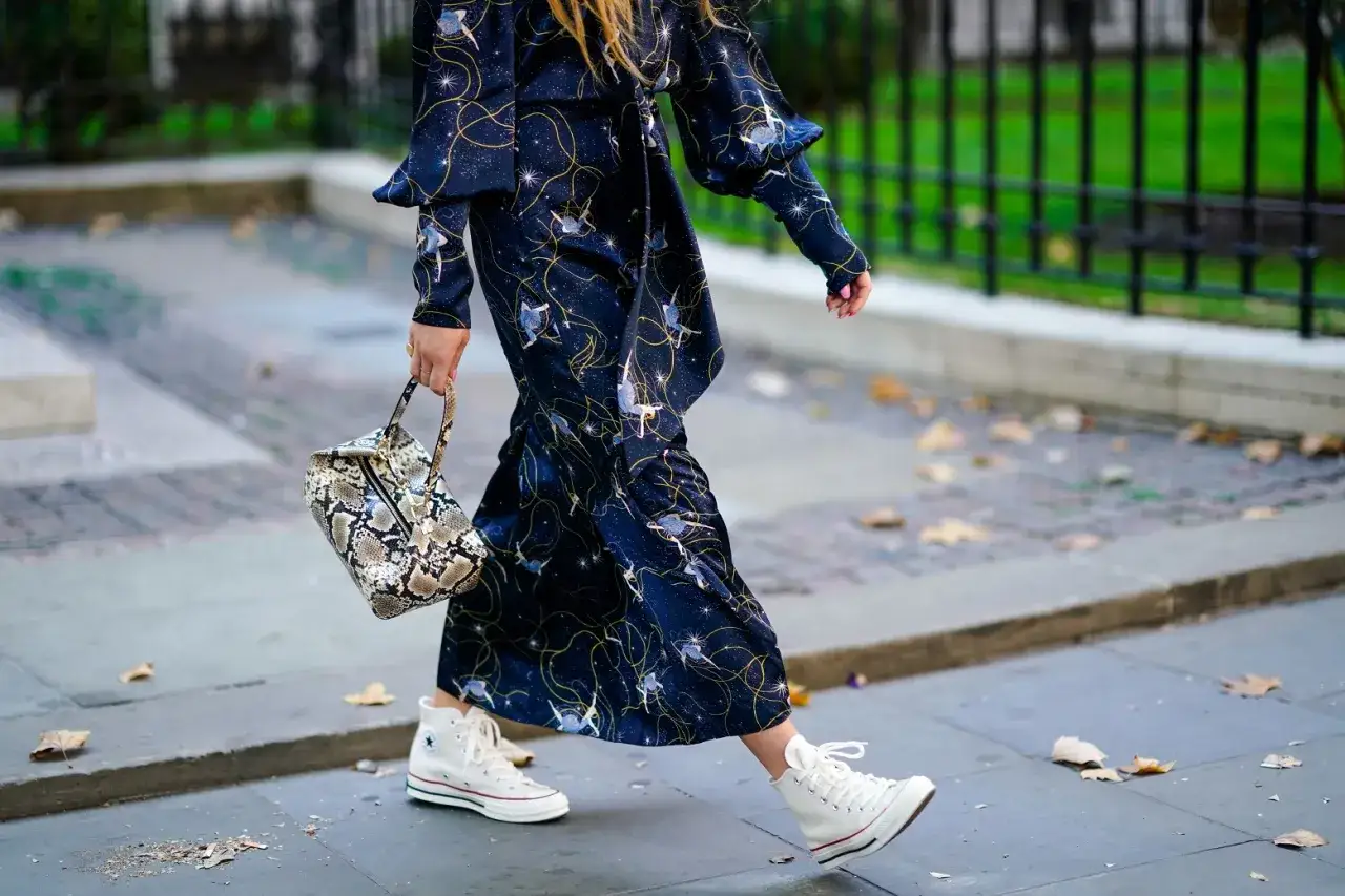 Kobieta w wieczorowej sukience w gwiazdy i księżyce, z torebką w panterkę, idzie w białych trampkach.