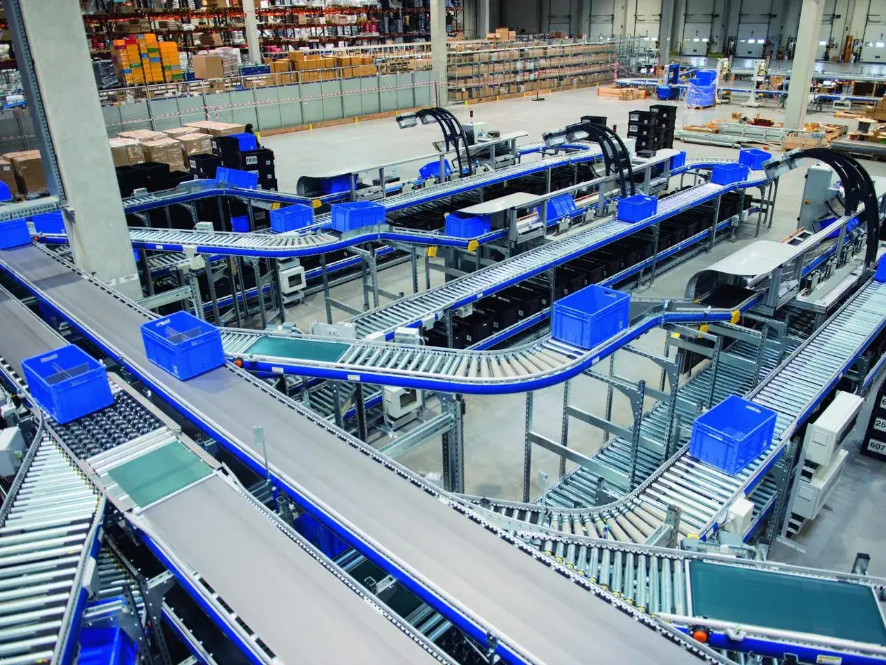 Automatyczne linie transportowe w magazynie, z niebieskimi skrzynkami przemieszczającymi się po taśmociągach. To pokazuje zaawansowane funkcje magazyn&oacute;w.