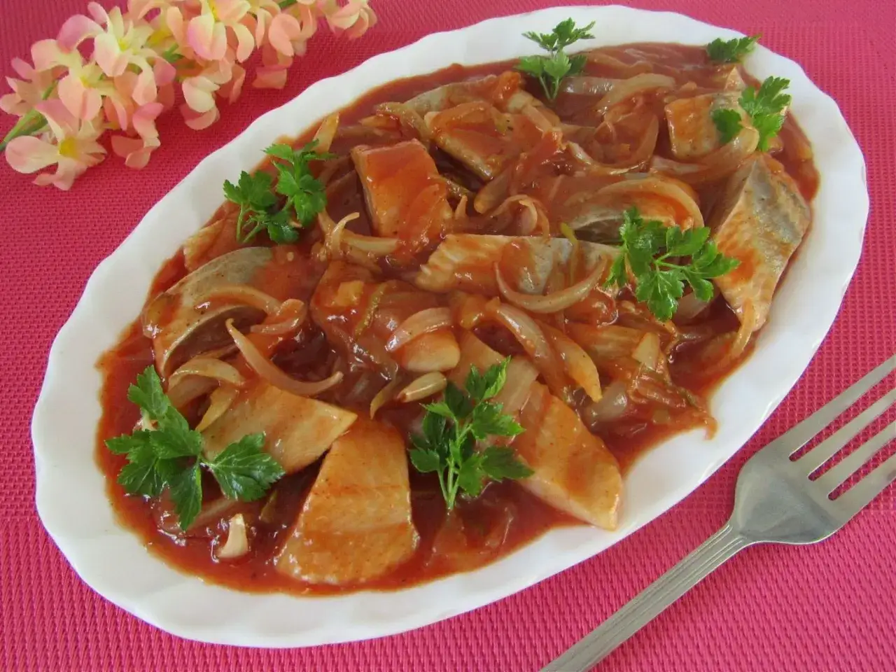 Śledzie po cygańsku na talerzu, w sosie pomidorowym z cebulą, ozdobione natką pietruszki. Pyszny przepis!