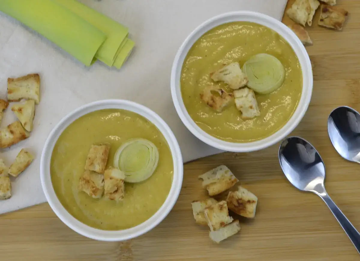 Dwie miseczki kremowej zupy z porów, udekorowane grzankami i plasterkiem pora. Obok świeże pory i łyżki.