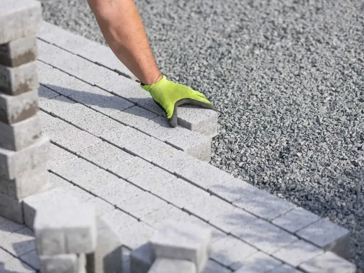 Ręka w zielonej rękawicy układa kostkę brukową na podsypce piaskowo cementowej.