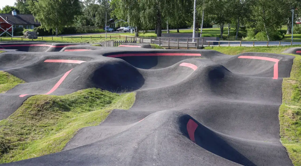 Asfaltowy pump track z czerwonymi pasami, otoczony zielenią i drzewami. Idealne miejsce do zabawy na rowerze lub hulajnodze.