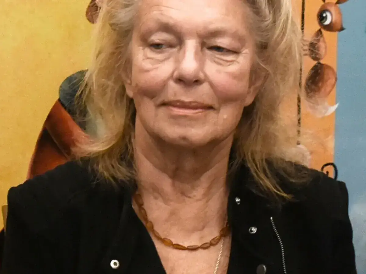 Katarzyna Gaertner, zamyślona, w czarnej kurtce i bursztynowym naszyjniku. W tle kolorowe obrazy, być może nawiązujące do jej dzieci.