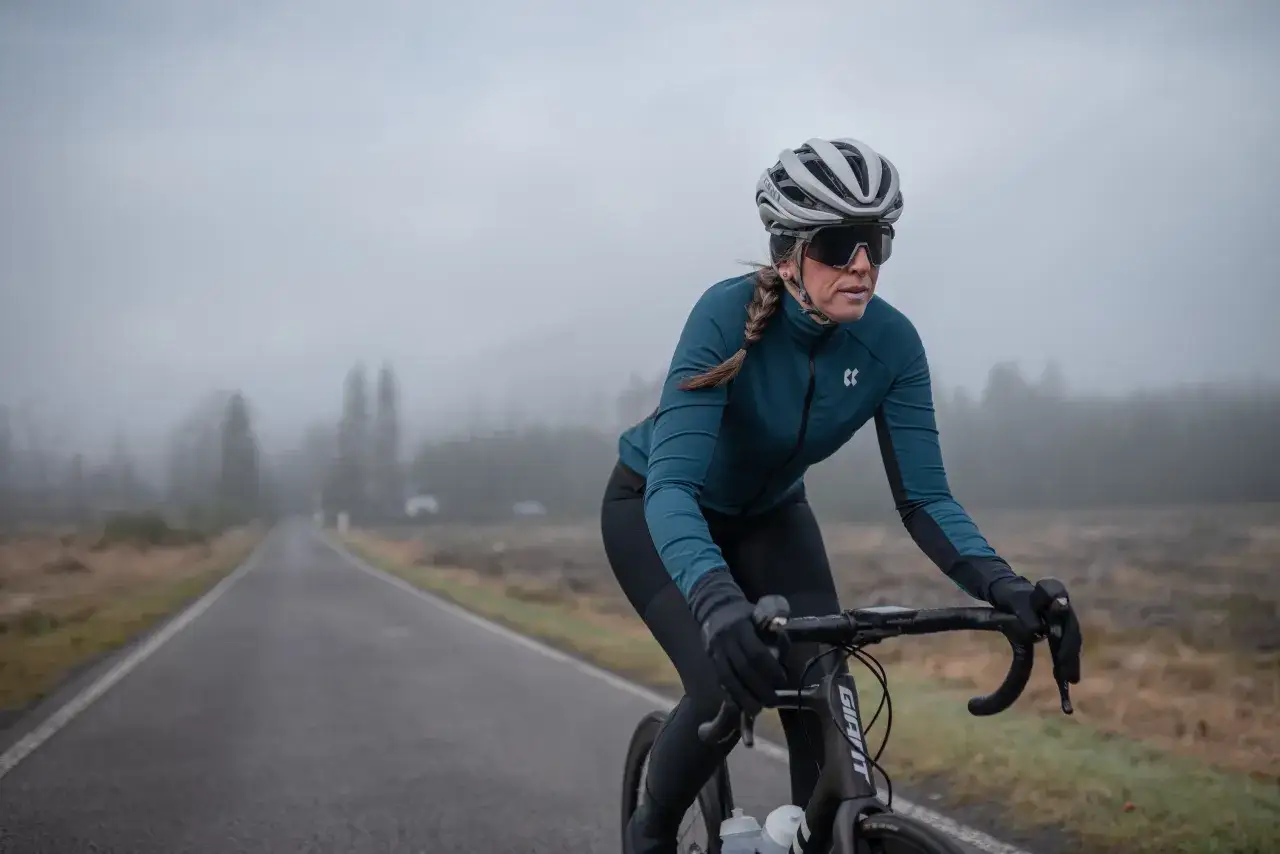Kobieta w kasku i okularach rowerowych jedzie na rowerze szosowym. Odpowiednie ubranie na rower to klucz do komfortu w chłodny, mglisty dzień.