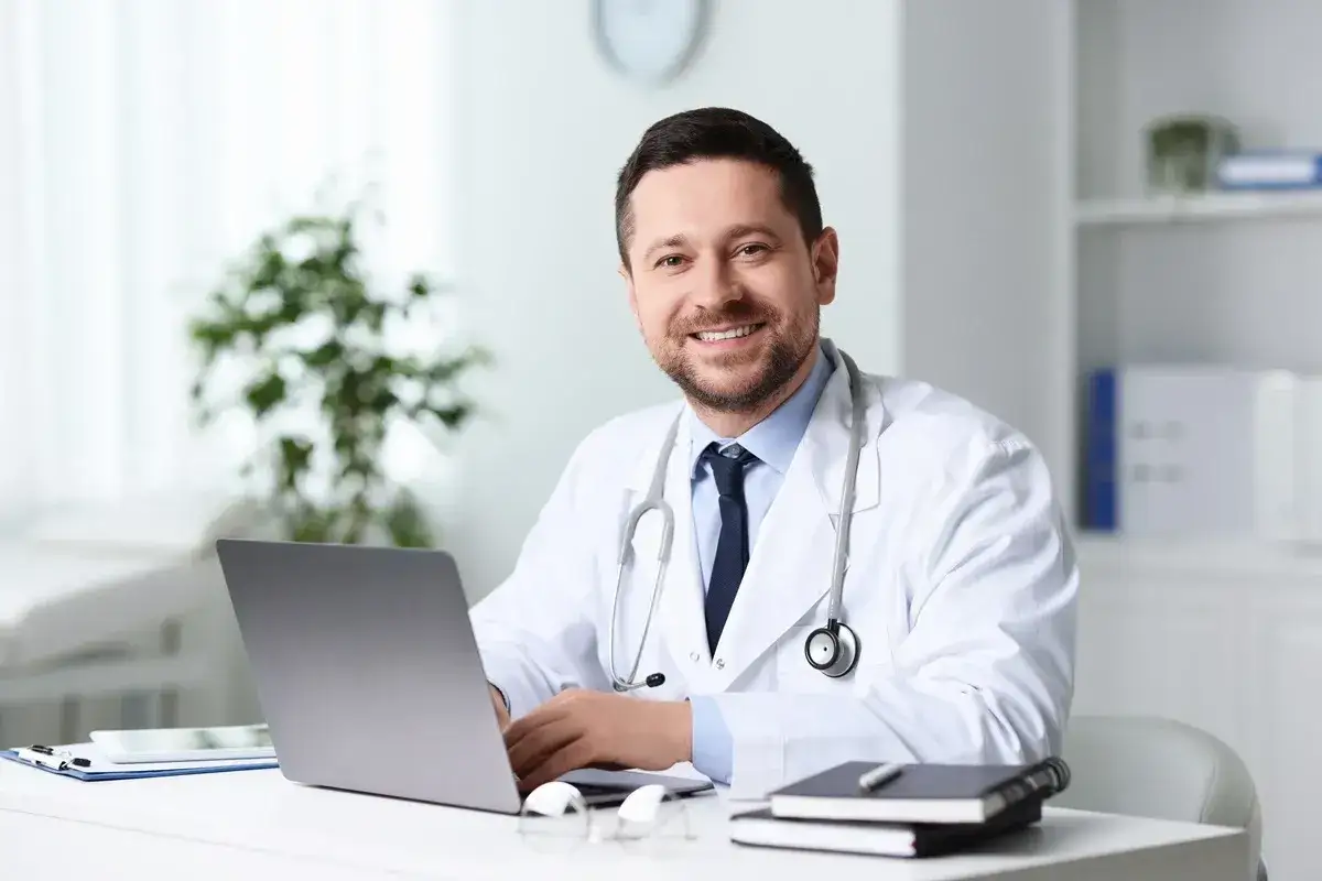 Uśmiechnięty lekarz przy biurku z laptopem. Pytanie o to, ile dni do przodu można wystawić L4, jest kluczowe dla pacjentów.