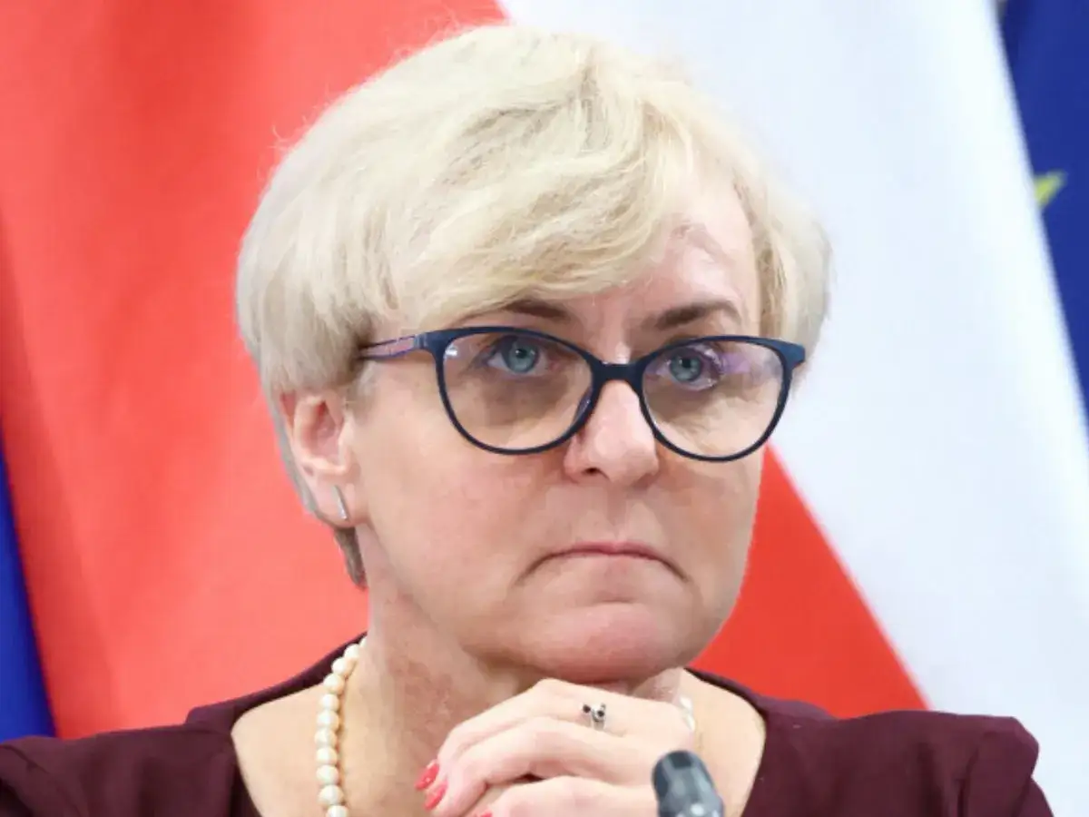 Joanna Kluzik Rostkowska w okularach, z rękami pod brodą, na tle flagi Polski i UE.