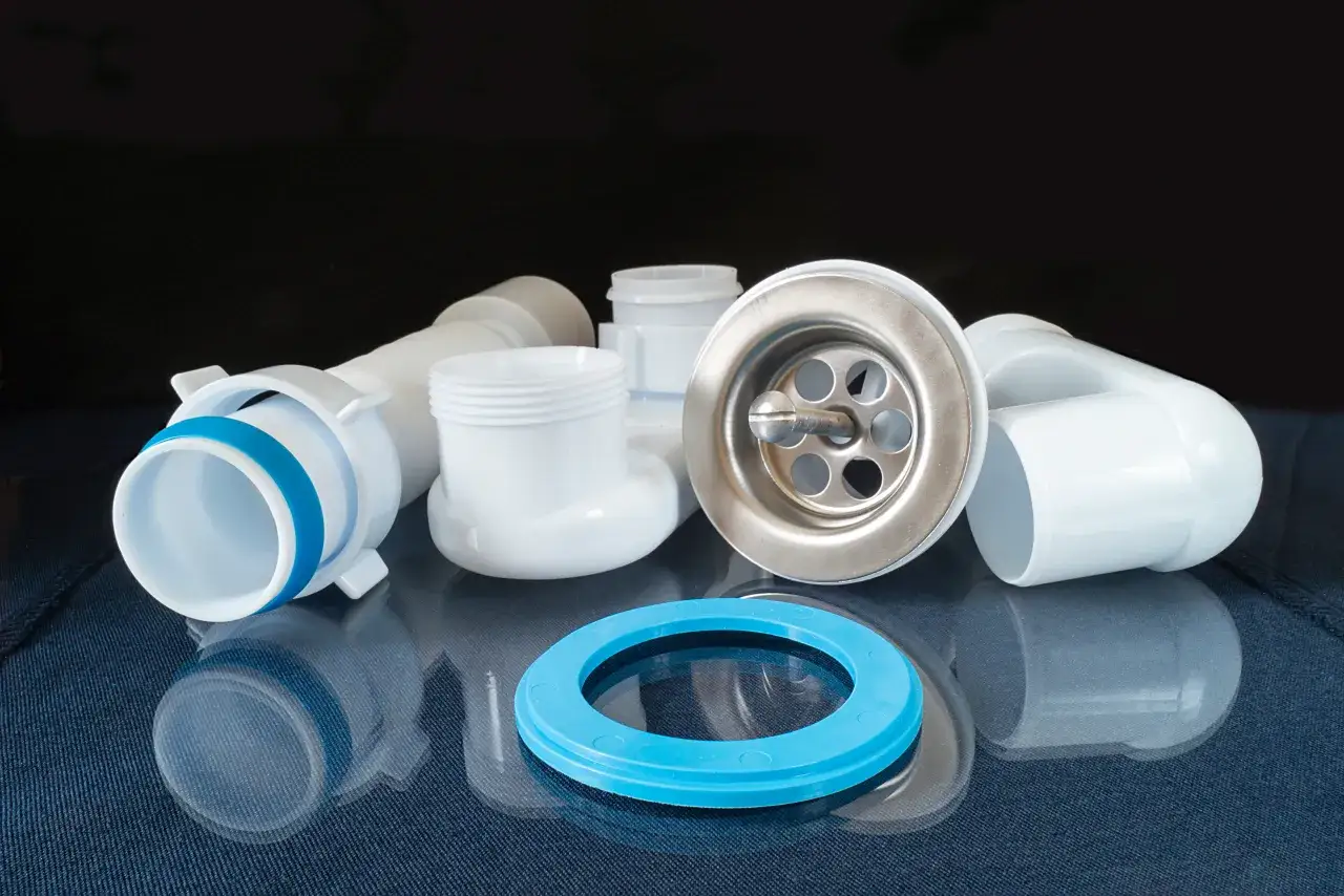 Białe elementy syfonu pod umywalkę, w tym metalowa nakrętka z sitkiem i niebieska uszczelka, leżą na granatowym tle.