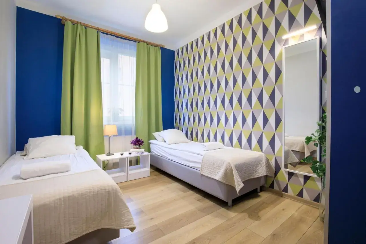 Pokój w nowoczesnym hostelu lub hotelu z dwoma łóżkami, zielonymi zasłonami i geometryczną tapetą.