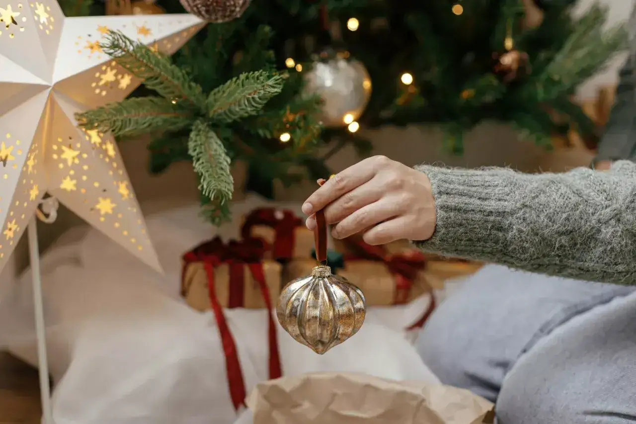 Ręka z ozdobą świąteczną pod choinką, kiedy ściągamy ozdoby świąteczne. W tle prezenty i gwiazda.