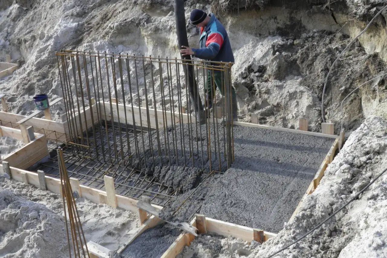 Robotnik wylewa beton do szalunku zbrojeniowego. Trwa praca nad fundamentami, a czas schnięcia betonu jest kluczowy.