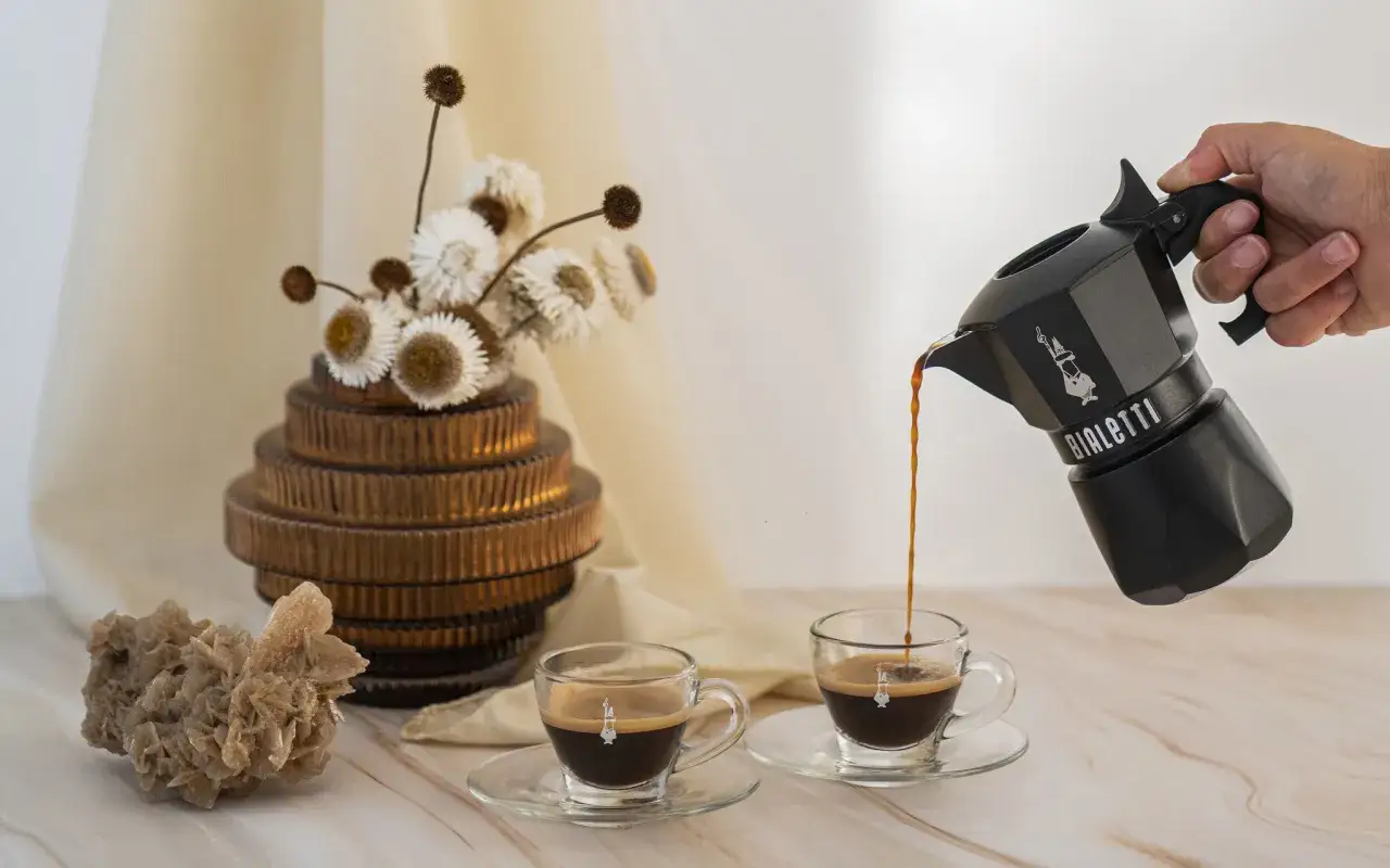 Aromatyczne kawy włoskie z kawiarki Bialetti nalewane do filiżanek. Obok dekoracje: wazon z suszonymi kwiatami i pustynna r&oacute;ża.