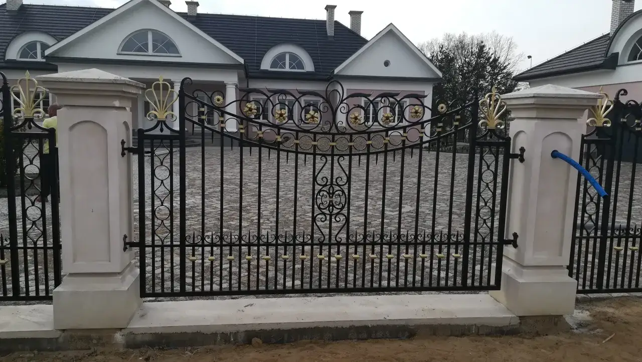 Elegancka brama kuta wzory z ozdobnymi złotymi elementami kwiatowymi i liśćmi, osadzona między kamiennymi słupami.