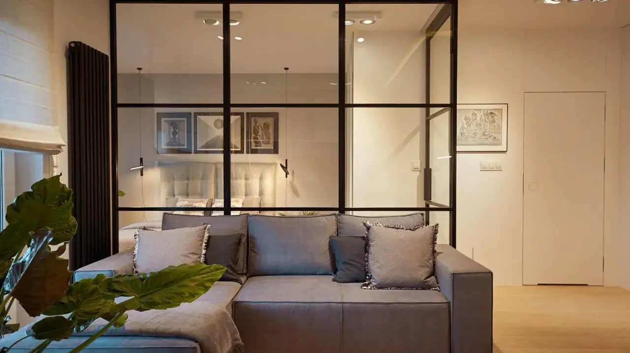Nowoczesny projekt mieszkania 45m2. Szara kanapa, szklana ściana z widokiem na sypialnię, duża roślina.