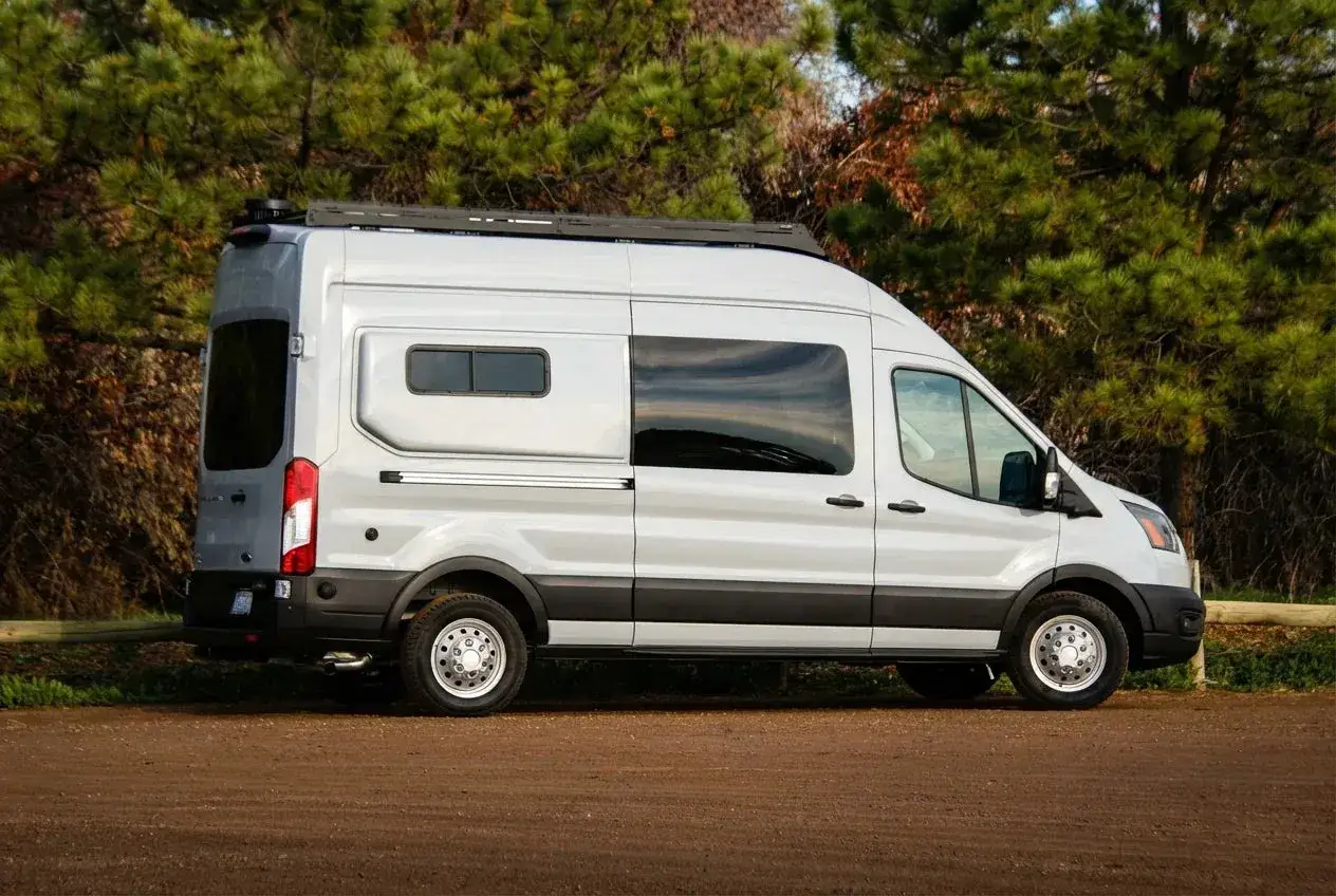 Nowy camper wypożyczenie gotowy na przygodę. Srebrny van z oknami i bagażnikiem dachowym, zaparkowany na drodze leśnej.