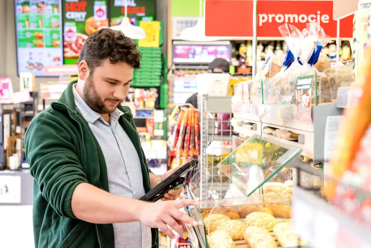 Pracownik Żabki sprawdza na tablecie, ile zarabia, przeglądając ofertę promocyjną i świeże pieczywo.