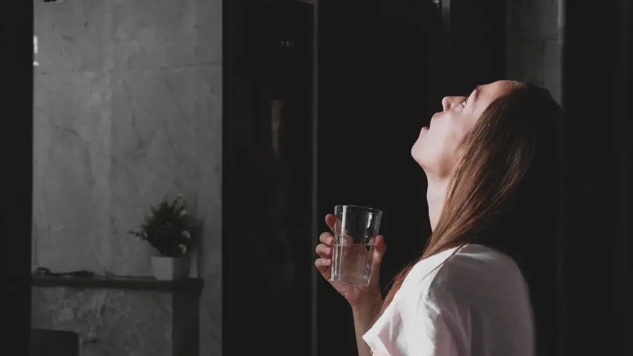 Kobieta pochyla głowę do tyłu, przygotowując się do płukania gardła solą. W dłoni trzyma szklankę z wodą.