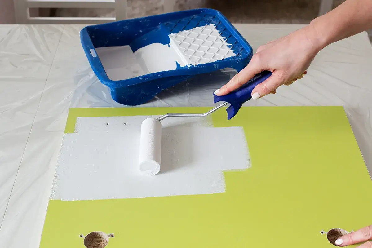 Malowanie frontu szafy z płyty na biało. Wałkiem nakładana jest farba, przygotowując mebel do odnowienia.