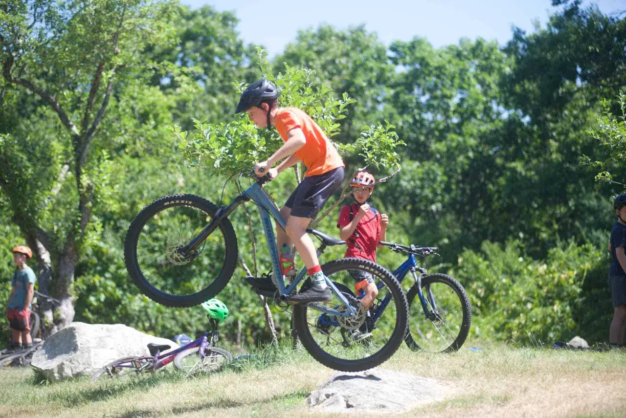 Chłopiec na rowerze MTB wykonuje skok. W tle inny rowerzysta i zielone drzewa.