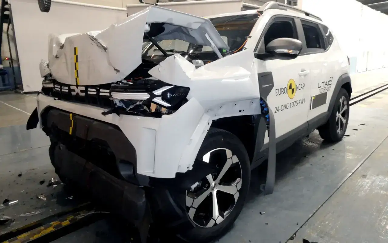 Biała Dacia Duster po teście zderzeniowym Euro NCAP. Przód samochodu jest mocno zniszczony, maska wygięta, a zderzak rozbity.