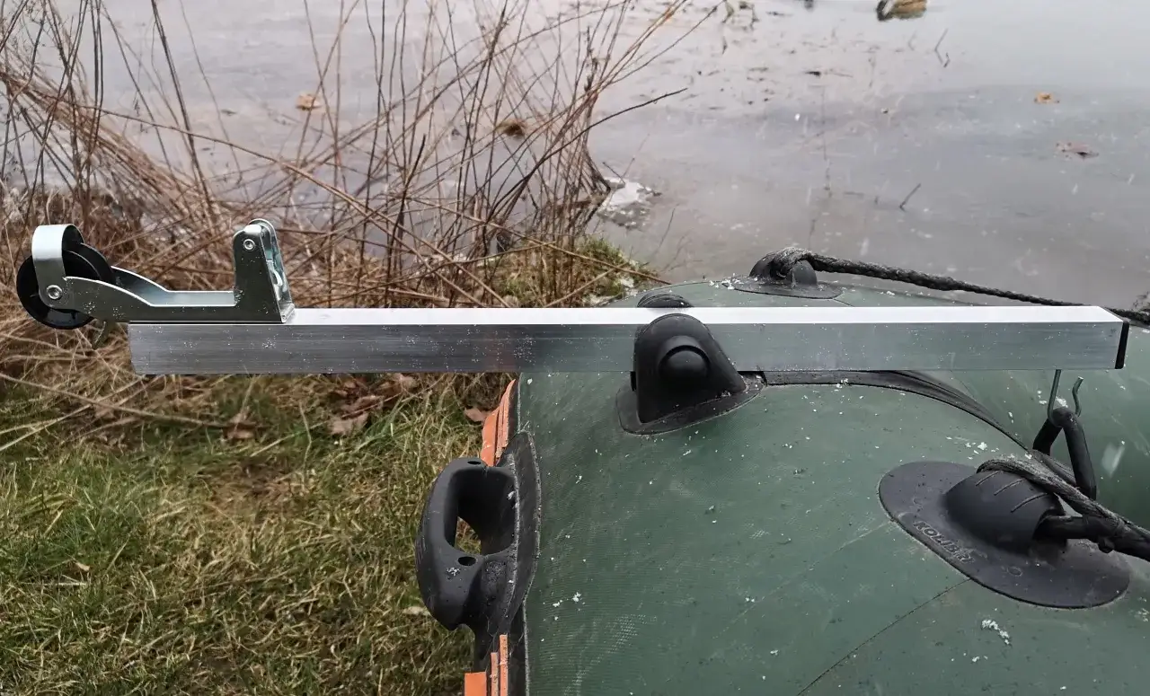 Metalowa kotwica do pontonu z kółkiem, zamontowana na zielonym pontonie przy brzegu porośniętym trawą i suchymi gałęziami.