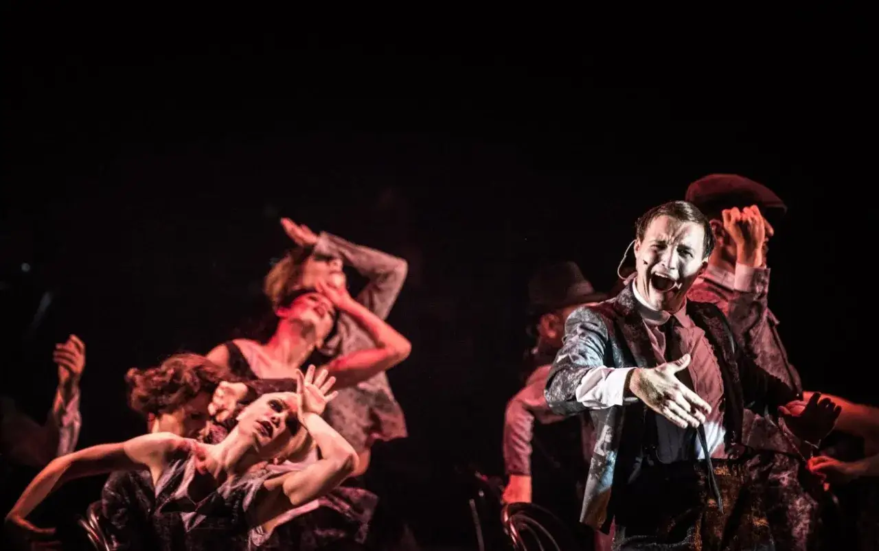 Kamil Krupicz w teatralnym szale, otoczony przez tancerzy w mrocznej scenerii.
